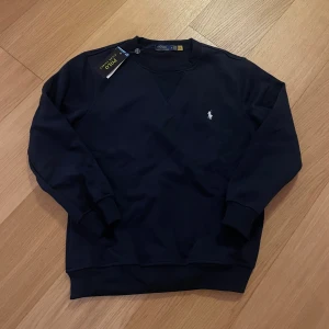 Mörkblå sweatshirt från Polo Ralph Lauren - Snygg mörkblå sweatshirt från Polo Ralph Lauren med klassisk rund hals och ribbade muddar. Tröjan har det ikoniska broderade logot på bröstet i vitt och är tillverkad i mjuk bomull. Perfekt för en chill och stilren look.