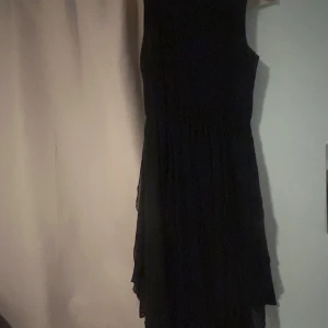 Svart kortklänning från H&M - Svart ärmlös lite längre klänning från H&M med lätt vidd i kjolen och markerad midja. Klänningen har ett tunt, flowy yttertyg och ett mjukt innertyg som gör den bekväm att bära. Perfekt för dig som gillar en enkel men stilren look.