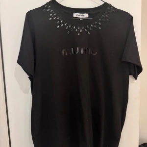 Svart t-shirt med stenar från Miu Miu - Svart t-shirt från Miu Miu med glansiga svarta stenar runt halsringningen och diskret logga framtill. Klassisk passform och korta ärmar. Tillverkad i mjuk bomull för en skön känsla. Perfekt för dig som vill ha en lyxig och stilren look. Storlek xl men uppskattar den till en m/l. Jag har användt den två ggr och märket sitter därför inte längre på men jag har kvar det. Köpt i Danmark för 900 DKK äkta 