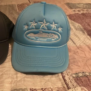 Två truckerkepsar med broderat skepp - Säljer två snygga truckerkepsar, en i ljusblått och en i svart. Båda har broderat motiv med ett skepp och fem stjärnor framtill. Kepsarna har böjd skärm och mesh på sidorna för extra ventilation. Perfekt accessoar för en cool streetstil.