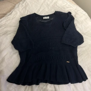 Mörkblå stickad blus från Holly & Whyte - Supersöt mörkblå stickad blus från Holly & Whyte med snyggt hålmönster, volangkant nedtill och små volanger vid axlarna. Blusen har rund hals och trekvartsärm, perfekt för dig som gillar detaljer och en feminin vibe.