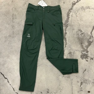 Gröna Haglöfs byxor - Gröna friluftsbyxor från Haglöfs i storlek M. Byxorna har flera praktiska fickor, bland annat en med dragkedja och en med knappstängning på benen. Justerbara benslut med kardborreband för bättre passform. Tillverkade i ett lätt och slitstarkt material som passar för vandring och utomhusaktiviteter.