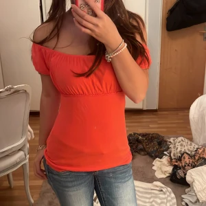 Orange röd offshoulder topp Vero Moda XS - Snygg orange topp från Vero Moda i storlek XS. Modellen är offshoulder med korta puffärmar och har en smickrande, figurnära passform. Tillverkad i mjuk bomull, perfekt för dig som gillar färgstarka och trendiga plagg. Elastisk söm under bysten för extra komfort.