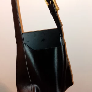Svart axelväska i skinn med guldspänne - Stilren svart axelväska i slätt skinn med justerbart axelband och snyggt guldspänne. Väskan har en enkel, fyrkantig form och rymmer enkelt en laptop eller böcker. Perfekt för dig som gillar minimalistisk och klassisk design.