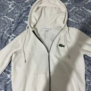 Vit zip tröja från Lacoste - Snygg vit hoodie från Lacoste i storlek M med klassisk krokodillogga på bröstet. Tröjan har huva med snörning, praktiska fickor och hel dragkedja framtill. Perfekt för dig som gillar stilrena och sportiga plagg.
