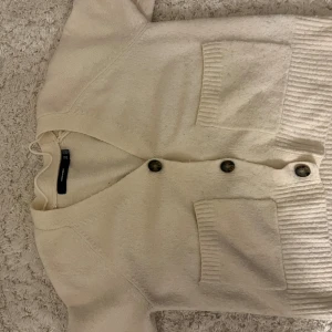 Beige kofta från Vero Moda - Mysig beige kofta från Vero Moda med ribbade detaljer och stora knappar framtill. Koftan har två praktiska fickor och en v-ringad hals. Perfekt att slänga på sig när det är lite kyligt ute. Passar dig som gillar enkel och stilren look.
