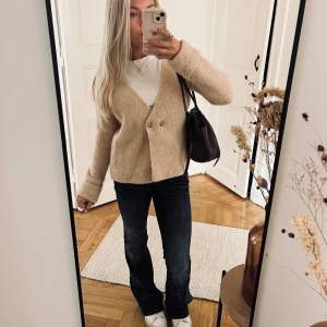 Beige stickad kofta från Zara - Supermysig beige stickad kofta från Zara med v-ringning och två knappar framtill. Koftan har lång ärm och ribbad struktur, perfekt för lager-på-lager. Snygg att styla med jeans eller kjol för en chill look.