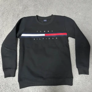 Svart Tommy Hilfiger sweatshirt L - Svart sweatshirt från Tommy Hilfiger i storlek L med klassisk logga och ränder i vitt, rött och mörkblått över bröstet. Tröjan har rund hals, långa ärmar och ribbade muddar. Materialet är mjuk bomull med inslag av polyester för extra komfort.