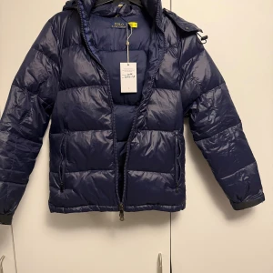 Mörkblå pufferjacka Polo Ralph Lauren - Snygg mörkblå pufferjacka från Polo Ralph Lauren med huva och dragkedja. Jackan har flera fickor med dragkedja och klassisk Polo-logga broderad på bröstet. Perfekt för kalla dagar och riktigt skön passform.