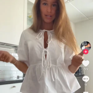 Vit blus med knyt och volang - Supersöt vit blus med två knyt framtill och lätt volang nedtill. Blusen har korta ärmar och en luftig passform som ger en romantisk vibe. Perfekt att styla med jeans eller kjol för en fräsch look.