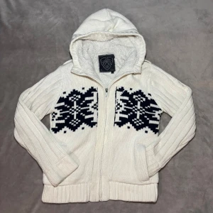 Stickad Fodrad Cardigan - Mysig vit stickad hoodie med svart vintermönster på bröstet. Tröjan har dragkedja, huva med fluffigt foder och ribbade muddar. Perfekt för kalla dagar när du vill vara både varm och snygg.