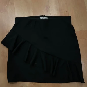 Svart kjol med volang från Skill - Medellång till kort kjol från Still med omlott-design och snygg volangdetalj på framsidan. Kjolen är i ett ribbat och stretchigt material som sitter bekvämt med normal passform. 