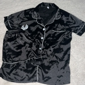 Svart satinpyjamas med vita detaljer - Snygg svart pyjamas i satin med kortärmad skjorta och matchande shorts. Skjortan har vita knappar, vit piping längs kanterna och en liten bröstficka. Materialet är glansigt och känns lyxigt mot huden – perfekt för chillkvällar hemma.