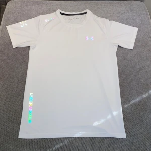 Vit Under Armour HeatGear t-shirt - Snygg vit t-shirt från Under Armour med reflekterande logga och textdetaljer. Tillverkad i lätt och snabbtorkande HeatGear-material som andas. Perfekt för träning eller sportiga dagar. Klassisk passform med korta ärmar och rund hals.
