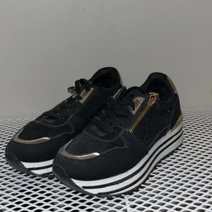 Svarta sneakers med guldiga detaljer - Snygga svarta sneakers med guldiga metalldetaljer och dragkedja på sidan. Skorna har en chunky plattformssula med svarta och vita ränder, rund tå och snörning framtill. Materialet är en mix av syntet och textil, vilket ger en cool och modern vibe.
