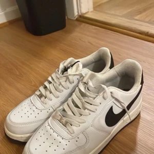 Nike Air Force 1 vita sneakers - Klassiska Nike Air Force 1 sneakers i vitt skinn med svart swoosh och svart yttersula. Skorna har snörning, perforerade detaljer på tån och metallbricka med 'AF1 82' på snörena. Perfekta för en stilren och sportig look.