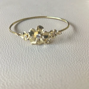 IOAKU la flour guld  - Ett aldrig använt Armband i guldfärg med tunnt band och stora blommor framtill. Blommorna har små glittrande stenar i mitten som ger extra detalj. Perfekt accessoar för att lyfta din stil med en feminin touch. 