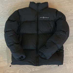 Svart Sail Racing pufferjacka - Svart pufferjacka från Sail Racing med hög krage och dragkedja framtill. Jackan är fylld med 700 fill power dun från Allied Feather + Down och har logotyp på bröstet samt en rund detalj på ryggen. Justerbar nederkant med snörning för extra komfort. Om du har frågor eller funderingar är det bara att höra av sig!