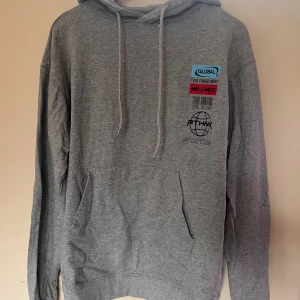 Grå hoodie från Rethink Status - Grå hoodie från Rethink Status med tryck och broderade detaljer på bröstet. Klassisk känguruficka, dragsko i huvan och mjukt fleecetyg på insidan. Perfekt för chill dagar och streetstyle. Snygga färgdetaljer i blått, rött och svart på trycket.