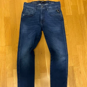 Replay Hyperflex mörkblå jeans - Säljer ett par Replay Hyperflex jeans i mörkblå tvätt med snygga slitningar. Klassisk femficksmodell med stretchigt material för extra komfort. Diskret Replay-logga på fickan och läderpatch bak i midjan. Perfekta för dig som gillar stilrena och bekväma jeans.