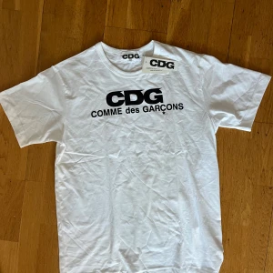 Vit t-shirt från Comme des Garçons - Snygg vit t-shirt från Comme des Garçons med stort svart CDG-tryck på bröstet. Klassisk rund hals och korta ärmar. Tillverkad i mjuk bomull som känns skön mot huden. Perfekt för dig som gillar streetwear och stilrena logotyper. 