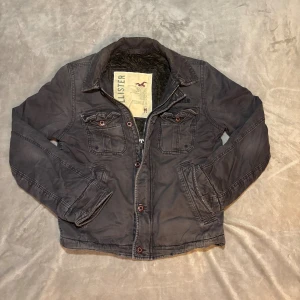 Mörkgrå jacka från Hollister - Snygg mörkgrå jacka från Hollister med två bröstfickor med knappar och dragkedja framtill. Jackan har mjukt foder på insidan och klassisk krage. Perfekt för kyliga dagar och har en casual look som funkar till det mesta.