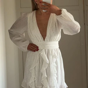 Vit omlottklänning från NBD med volanger - Supersöt vit kortklänning från NBD med djup v-ringning, långa puffärmar och omlottkänsla. Klänningen har markerad midja, volangdetaljer framtill och små silvriga detaljer i tyget. Perfekt till student. Nypris runt 3500