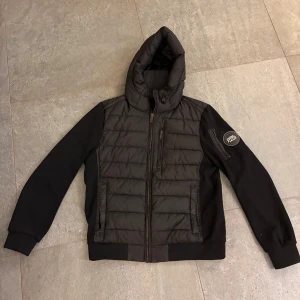 Svart puffer cardigan Supply & Demand L - Svart pufferjacka från Supply & Demand med quiltad front och huva. Jackan har dragkedja, ribbade muddar och snygg logotyp på ärmen. Materialmix med polyester och syntet, perfekt för kyligare dagar. Stilsäker och urban look.