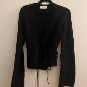 Svart omlottkofta från Gina Tricot - Svart omlottkofta från Gina Tricot i ullblandning. Koftan har långa ärmar, knytband i midjan och en mysig, lite luddig yta. Perfekt för lager-på-lager och snygg till både jeans och kjol.