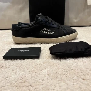 Svarta Saint Laurent sneakers - Säljer ett par svarta Saint Laurent sneakers med handskriven logga på sidan. Skorna har snörning, rund tå och en beige platt gummisula. Ovandelen är i canvas och skinn, med slitna detaljer för en cool look. Kommer med originalkartong och dustbags. Såklart riktiga och kostar runt 7000 nya
