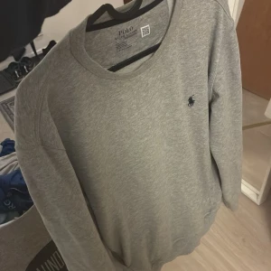 Grå sweatshirt från Polo Ralph Lauren - En stilren grå sweatshirt från Polo Ralph Lauren med klassisk rund halsringning och broderad logga på bröstet. Tröjan har långa ärmar och praktisk ficka med dragkedja på sidan. Perfekt för chill dagar eller när du vill vara casual. Lite stor i storleken så passar m också