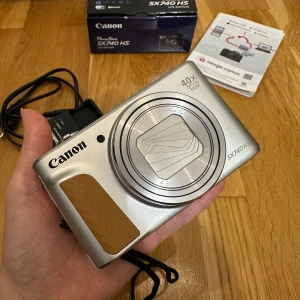 Canon PowerShot SX740 HS - Canon PowerShot SX740 HS digitalkamera i silver med 40x optisk zoom, 20.3 MP sensor, 4K-video och WiFi. Kameran är i mycket fint skick, inga synliga repor eller skador. Den är inköpt i maj 2025 och har endast används under en kort resa. Garanti finns kvar. Levereras med originalkartong, laddare, batterr och manual. Den kommer även med ett extra batteri, ett glasskydd som sitter på och ett skal.