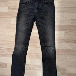 Svarta jeans från Nudie Jeans W27 L30 - Snygga svarta jeans från Nudie Jeans med klassisk femficksdesign och coola slitningar framtill. Jeansen har raka ben och mörk tvätt med Nudie-loggan broderad på bakfickorna. Perfekt för dig som gillar stilrena och tidlösa jeans. I nyskick 