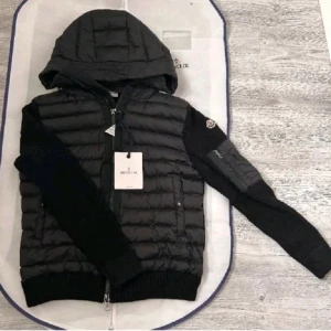 Svart Moncler cardiganjacka med huva - Svart cardiganjacka från Moncler med stickade ärmar och quiltad kropp. Jackan har en stor huva, dragkedja framtill och ribbade muddar. Märkeslogga på ärmen och praktiska fickor med knappstängning. Perfekt för dig som gillar stilren och sportig look.Skriv vilken storlek du har.
