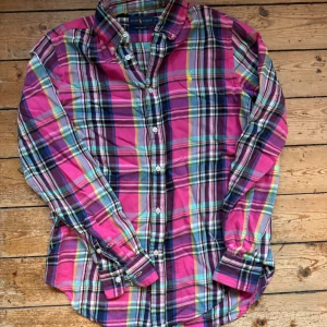 Rutig skjorta från Ralph Lauren - Färgglad rutig skjorta från Ralph Lauren i relaxed fit. Skjortan har en mix av rosa, blå, gul och gröna toner, klassisk krage och knäppning framtill. Tillverkad i mjuk bomull och prydd med den ikoniska broderade loggan på bröstet.