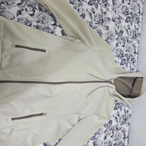 Beige vindjacka med huva och dragkedja - Snygg beige vindjacka med huva och justerbar snörning. Jackan har två praktiska fickor med dragkedja och hel dragkedja framtill. Materialet är mjukt och syntetiskt, perfekt för blåsiga dagar. Köpt för 799 kr