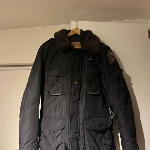 Svart vinterjacka från Parajumpers - Parkajacka från Parajumpers i svart med brunt teddyfoder på kragen och patch på ärmen. Jackan har flera fickor med lock, dragkedja och knappar framtill samt justerbar midja. Perfekt för kalla dagar och har en cool rescue squadron-vibe.