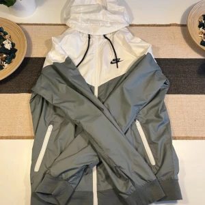 Nike vindjacka vit och grå S - Nike vindjacka i snygg vit och grå färg med huva och dragkedja framtill. Jackan har två sidofickor med dragkedja, svart Nike-logga på bröstet och svarta snören vid huvan. Tillverkad i lätt polyester med meshfoder för extra komfort.