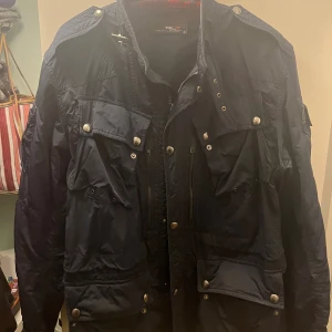 Mörkblå fieldjacka från polo Ralph Lauren - Snygg mörkblå RLX Ralph Lauren vindjacka med flera fickor, silverfärgade knappar och dragkedjor. Jackan har en cool militärinspirerad look och är tillverkad i lätt polyester. 