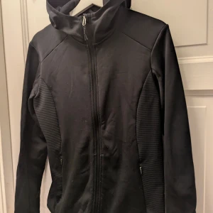 Svart sportig fleecejacka från H&M - Svart fleecejacka från H&M Sport med huva och hel dragkedja framtill. Jackan har ribbade paneler på sidorna, två fickor med dragkedja och långärmad design. Perfekt för aktiva dagar och har en clean, sportig look.