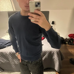 Mörkblå stickad tröja  - Säljer en stilren mörkblå stickad tröja  med diskret logga broderad på bröstet. Tröjan har rund hals, ribbade muddar och är långärmad. Perfekt för dig som gillar klassisk och enkel stil.