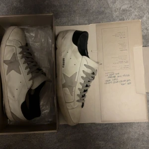 Golden Goose Superstar sneakers vit/grå - Golden Goose Superstar sneakers i vitt skinn med grå mockadetaljer och stjärna på sidan. Den är köpt här på Plick✅men tyvärr så är den i ganska tråkig/acceptabelt skick eftersom att nedre sulorna är slitna och gått upp❌. Jag rekommenderar att lämna de in till en skomakare. Pris kan diskuteras!!!