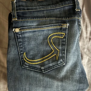 Rock & Republic blå bootcut jeans - Snygga blå jeans från Rock & Republic med bootcut passform. Jeansen har klassisk femficksdesign, coola kontrastsömmar i gult och tydlig broderad bakficka. Tillverkade i slitstarkt denim med en mörk tvätt och lätt slitning för en trendig look.