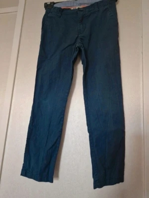 Mörkblå chinos i bomull - Snygga mörkblå chinos med klassisk rak passform och bälteshällor. Byxorna är tillverkade i mjuk bomull och har knappstängning framtill. Slitage syns på höger knä och det är en liten fläck vid grenen (se bilder). Möjligtvis kan färgen ha mattats av lite i och med tvätt. Stl 122