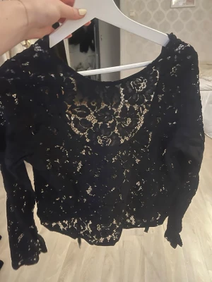 Svart spetsblus med blommönster - Säljer en svart långärmad blus i genombruten spets med blommigt mönster. Blusen har rund halsringning och är helt täckt av vacker spets som ger en elegant och trendig look. Perfekt att styla med jeans eller kjol för en snygg outfit.