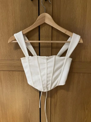 Vit korsett med snörning - Säljer en stilren vit croppad korsett med breda axelband och snygga sömdetaljer framtill. Baksidan har snörning som ger en cool och justerbar passform. Perfekt att styla över en skjorta eller t-shirt för en trendig look.