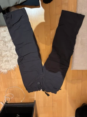 Svarta täckbyxor från The North Face - Säljer ett par svarta täckbyxor från The North Face med justerbar midja och stora fickor framtill. Byxorna har praktiska spännen och är gjorda i slitstarkt syntetmaterial, perfekta för kalla dagar i backen eller vinterpromenader.