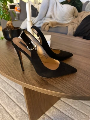 Svarta klackar med spetsig tå och hög klack - Säljer ett par stilrena svarta klackskor med spetsig tå och hög smal klack. Skorna har öppen häl och justerbart spänne runt ankeln. Materialet är mocka. Pris kan diskuteras 