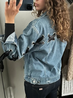 Jeansjacka med snörning och svarta band - Säljer en blå jeansjacka med unika svarta snörningsdetaljer längs ryggen och ärmarna. Jackan har klassisk krage, silverfärgade knappar framtill och en loose passform. Materialet är denim och jackan har coola detaljer som gör den till ett statement-plagg.
