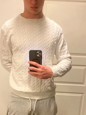 Vit kabelstickad tröja Cap Horn - Säljer en fräsch vit kabelstickad tröja från Cap Horn i ekologisk bomull. Tröjan har rund hals, ribbade muddar och klassiskt kabelmönster som ger en clean och stilren look. Perfekt till jeans eller chinos för en enkel men snygg outfit.